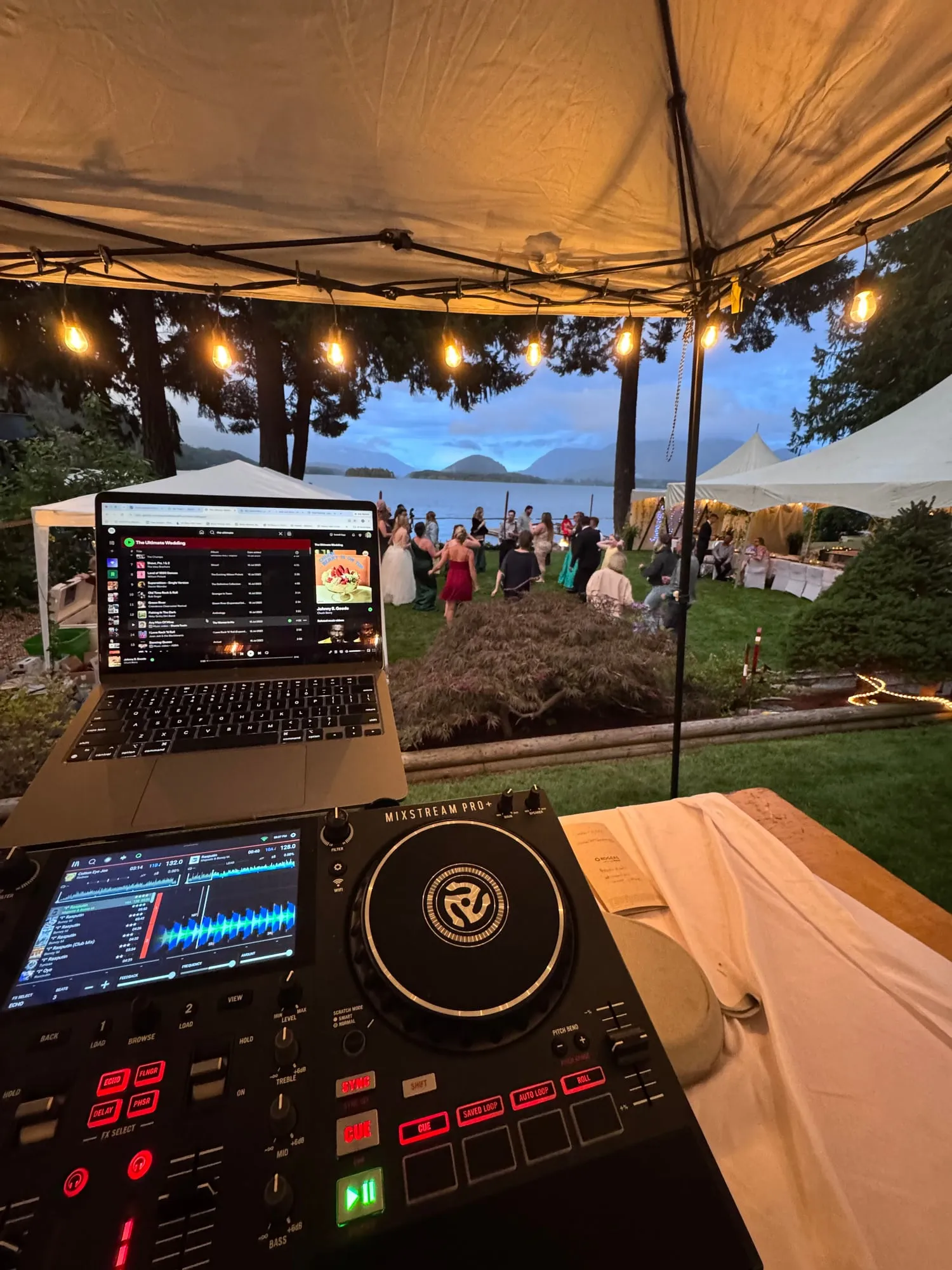 Ryan’s DJ controller and laptop under a string-lit tent at an outdoor wedding, guests dancing on the lawn beside the water at dusk